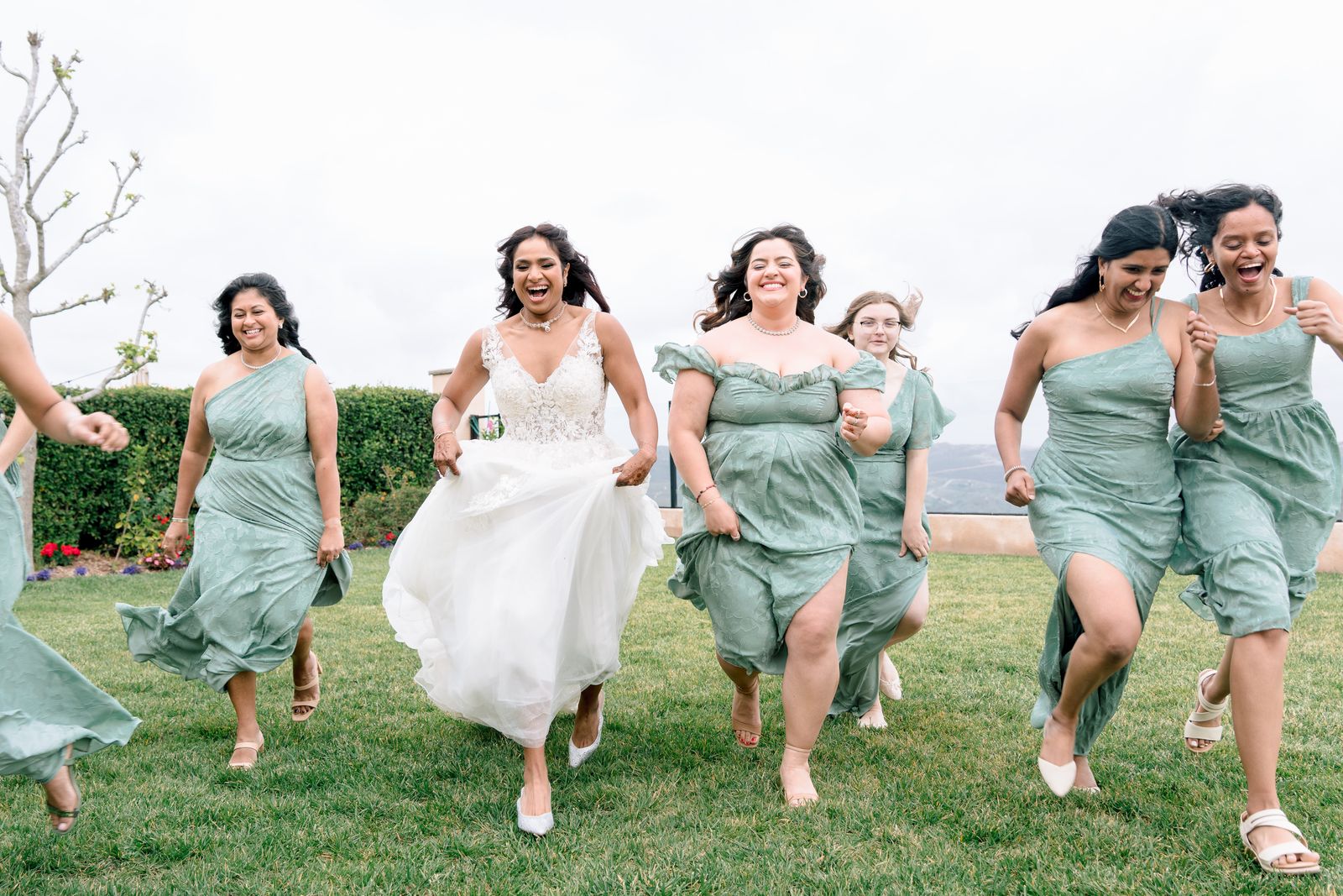 Bride and bridesmaids in sage dresses running and laughing across a lawn