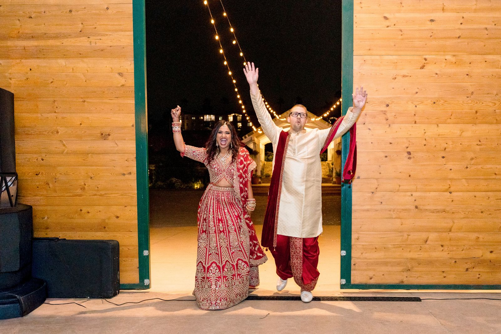 Couple entering their reception in a red lehenga and sherwani, arms raised